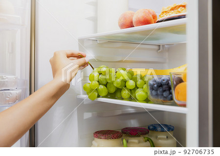 Woman hand taking, grabbing or picks up green bunch of grapes out of open refrigerator shelf or fridge drawer full of fruits, blueberries, bottles with yogurt. Healthy food diet, lifestyle concept 92706735