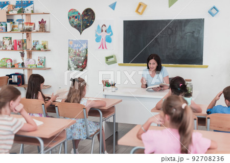 A teacher reads a book to elementary school students who listen carefully while sitting in a modern classroom A teacher reads a book to elementary school students who listen carefully while sitting in a modern classroom 92708236