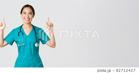 Covid-19, healthcare workers, pandemic concept. Smiling beautiful asian female nurse, doctor in scrubs pointing and looking up with satisfied grin, happy to show awesome promo offer, white background 92712427