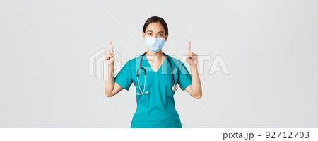 Covid-19, coronavirus disease, healthcare workers concept. Young professional asian female doctor, nurse in medical mask and scrubs, pointing fingers up, showing way, advertising 92712703