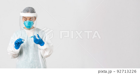 Covid-19, coronavirus disease, healthcare workers concept. Serious-looking female physician, asian doctor in personal protective equipment insert syringe in ampoule with vaccine, white background 92713226