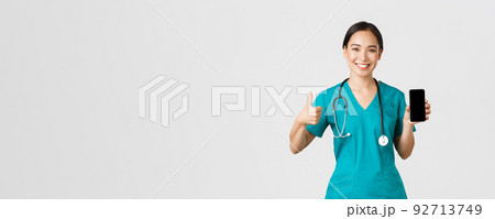 Covid-19, healthcare workers and online medicine concept. Portrait of young asian female physician, doctor in scrubs show thumbs-up and smartphone screen, promote application, white background 92713749