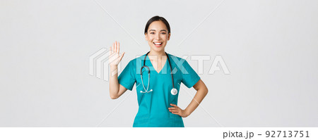 Covid-19, healthcare workers and preventing virus concept. Friendly-looking smiling asian female doctor, physician in scrubs waving hand to say hi, hello, greeting patient, nice to see you 92713751