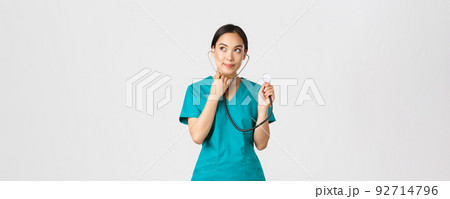 Covid-19, healthcare workers and preventing virus concept. Cute smiling asian female intern learn how listen lungs with stethscope, doctor examine patient, looking thoughtful upper left corner 92714796