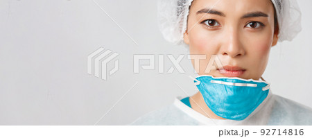 Covid-19, coronavirus disease, healthcare workers concept. Close-up of exhausted serious-looking asian female doctor take-off personal protective equipment, have skin damage from respirator 92714816