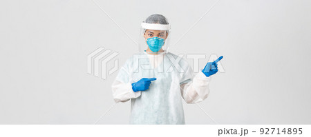 Covid-19, coronavirus disease, healthcare workers concept. Professional female doctor, tech lab employee in personal protective equipment pointing fingers right, showing way, white background Covid-19, coronavirus disease, healthcare workers concept. Professional female doctor, tech lab employee in personal protective equipment pointing fingers right, showing way, white background 92714895