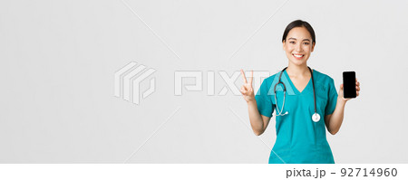 Covid-19, healthcare workers and online medicine concept. Smiling confident asian female doctor, physician in scrubs show peace sign and mobile screen, promote consultation app, white background 92714960