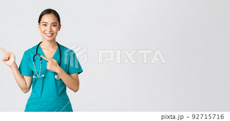 Covid-19, healthcare workers, pandemic concept. Smiling pretty asian doctor, nurse in scrubs and stethoscope, pointing fingers upper left corner, showing online clinic banner, white background 92715716