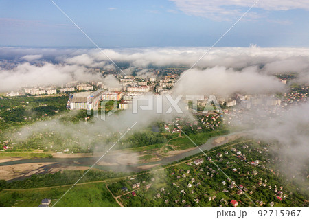 Aerial view of white clouds above a town or village with rows of buildings and curvy streets between green fields in summer. Countryside landscape from above. 92715967