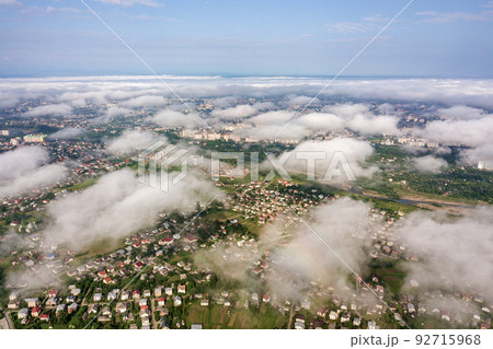 Aerial view of white clouds above a town or village with rows of buildings and curvy streets between green fields in summer. Countryside landscape from above. Aerial view of white clouds above a town or village with rows of buildings and curvy streets between green fields in summer. Countryside landscape from above. 92715968