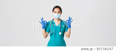Covid-19, coronavirus disease, healthcare workers concept. Smiling professional and confident asian doctor, physician in medical mask, gloves and scrubs show okay gesture, guarantee quality service 92716247
