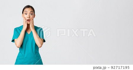 Covid-19, healthcare workers, pandemic concept. Surprised and startled asian nurse, doctor in scrubs stare at camera speechless, gasping amazed, listen to announcement, being impressed 92717195