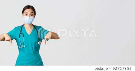 Covid-19, coronavirus disease, healthcare workers concept. Excited and surprised asian female physician, nurse in scrubs and medical mask, pointing fingers down, showing advertisement 92717352