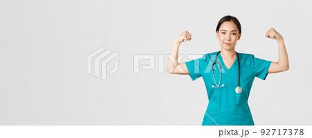 Covid-19, healthcare workers and preventing virus concept. Confident professional asian female doctor, nurse in scrubs smiling sassy and flex biceps, being strong, show-off muscles 92717378
