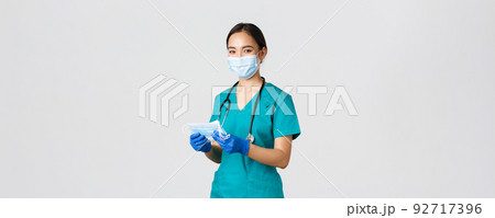Covid-19, coronavirus disease, healthcare workers concept. Smiling friendly female nurse, physician in scrubs and rubber gloves provide patients with medical masks, white background 92717396