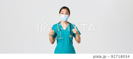 Covid-19, coronavirus disease, healthcare workers concept. Impressed happy asian female doctor, physician in medical mask and scrubs show thumbs-up, agree, compliment choice, praise nice work 92718034