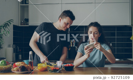 Attractive couple in the kitchen at home. Curly girl playing video game on smartphone while her boylfriend cooking breakfast 92725109