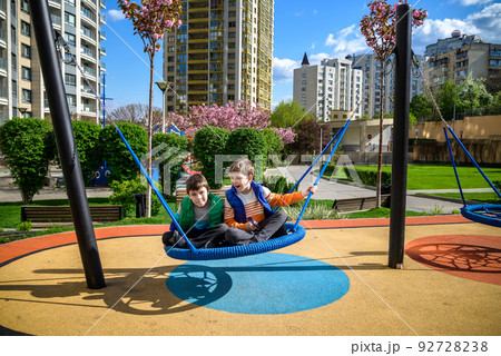 Cheerful boy with his toddler brother swinging in the swing-nest. Entertainment for preschoolers outdoors. Relaxation friendship concept Cheerful boy with his toddler brother swinging in the swing-nest. Entertainment for preschoolers outdoors. Relaxation friendship concept 92728238