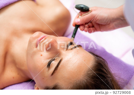 Relaxing massage. Side view european woman getting massage with jade face roller gouache in spa salon 92729008