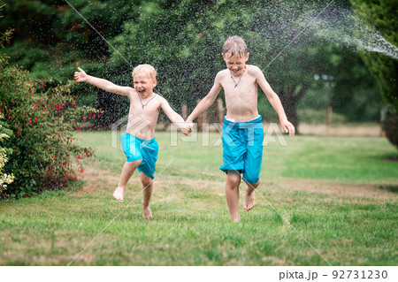 Kids play with water on hot summer day. Children with garden sprinkler. Outdoor fun. Boys run on a field under water drops. 92731230
