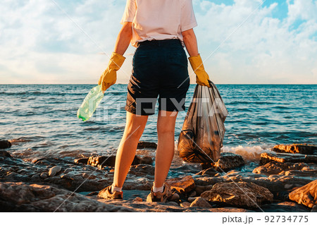 A woman stands with a plastic bag and bottle in her hands. Rear view. Close up of legs. Copy space. Cleaning of the coastal zone. The concept of Earth Day 92734775
