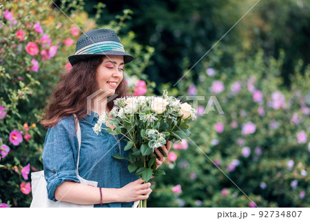 Portrait of a young happy woman in a straw hat holding a bouquet of white roses. Flowering bushes in the background. The concept of March 8 and Mother's Day Portrait of a young happy woman in a straw hat holding a bouquet of white roses. Flowering bushes in the background. The concept of March 8 and Mother's Day 92734807
