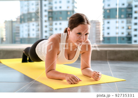 Girl in sportswear trains, doing plank on elbows, on fitness mat on balcony of house. 92735331