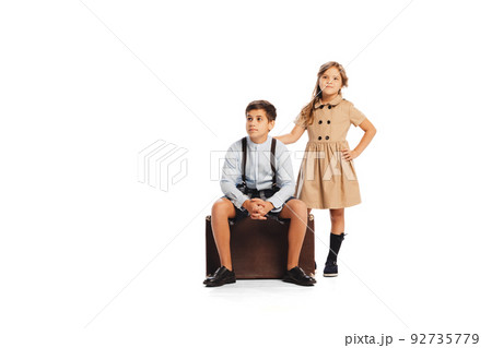 Portrait of stylish children in retro styled outfits posing, sitting on retro suitcase isolated over white studio background. Travelling 92735779