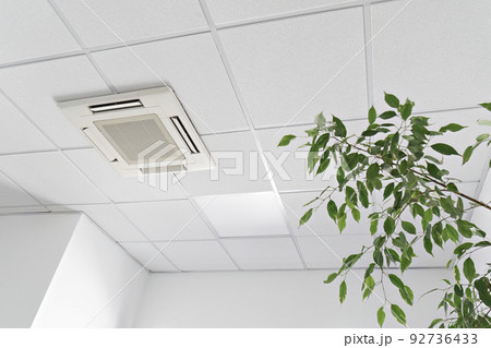 Low angle of assette Air Conditioner on ceiling in modern light office or apartment with green ficus plant leaves. Indoor air quality 92736433