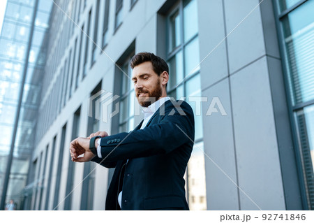 Smiling confident handsome young european male with beard in suit looks at watch, waits client Smiling confident handsome young european male with beard in suit looks at watch, waits client 92741846