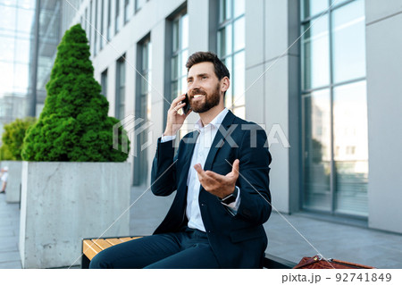 Happy millennial attractive european man with beard in suit calling by phone to client Happy millennial attractive european man with beard in suit calling by phone to client 92741849