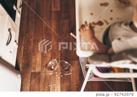 A small baby on a high chair dropped a cup of noodle soup on the wooden floor. The transparent plate is divided into two parts. Noodles in the form of figures. The table is dirty. High quality photo 92742079