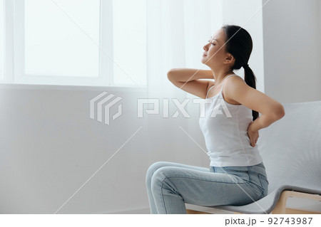 Back pain. Suffering from osteochondrosis after long study work pretty young Asian woman touching painful lower back at home interior living room. Injuries Poor health Illness concept. Cool offer 92743987