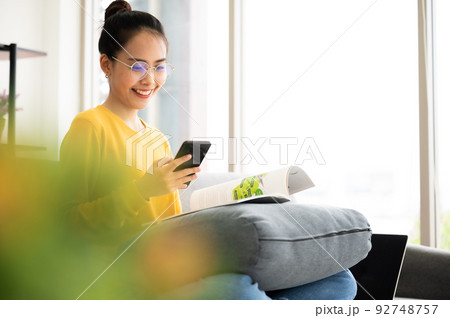 Happy young asian woman reading on the sofa and holding mobile smartphone in living room at home Happy young asian woman reading on the sofa and holding mobile smartphone in living room at home 92748757