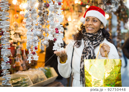 Cheerful Latina in Santa hat looking for New Year decorations in street market Cheerful Latina in Santa hat looking for New Year decorations in street market 92750448