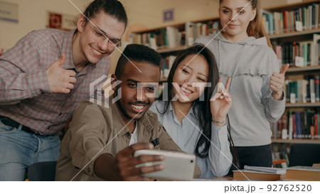 Group of international students have fun smiling and making selfie photos on smartphone camera at university library indoors. Cheerful friends have rest while preapre project together Group of international students have fun smiling and making selfie photos on smartphone camera at university library indoors. Cheerful friends have rest while preapre project together 92762520