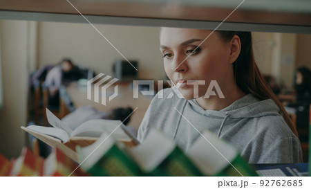 Young caucasian beautiful female student is standing near shelf with books in big lighty spacious library holding one, turning over pages and reading seriously 92762685