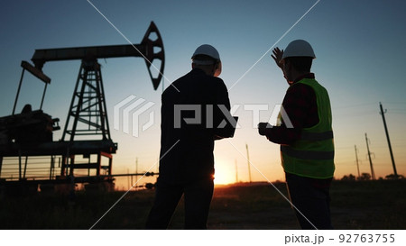 oil production. two workers a work next to an oil pump at sunset silhouette. industry business oil production concept. engineers lifestyle in hardhats studying oil production figures 92763755