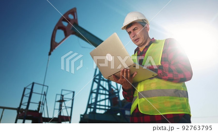 oil industry. engineer next to an oil rig. oil production business finance concept. gas production. a worker with a laptop monitors oil production next to an station. a worker sun in hard hat works 92763795