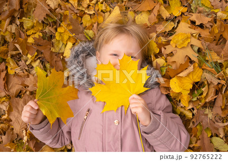 Adorable toddler blond girl lieing on the ground and playing with yellow fallen leaves in autumn park. 92765222
