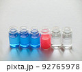 白い画用紙の上に横に並べた青色と赤色と透明な液体が入った6個のガラス瓶 92765978