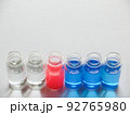 白い画用紙の上に横に並べた青色と赤色と透明な液体が入った6個のガラス瓶 92765980