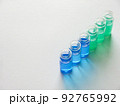 白い画用紙の上に斜めに並べた青色と緑色の液体が入った6個のガラス瓶 92765992