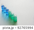 白い画用紙の上に斜めに並べた青色と緑色の液体が入った6個のガラス瓶 92765994