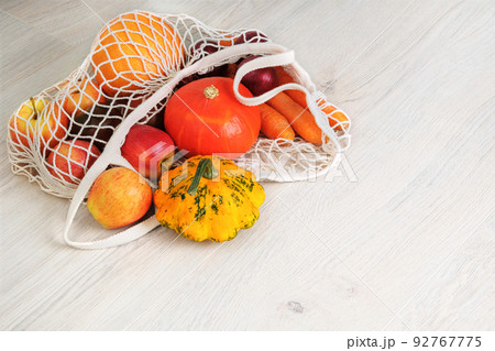 Orange pumpkin, apples, onions, zucchini and carrots in natural reusable mesh bag. Organic vegetables coming out from eco shopping bag. White wooden background. 92767775