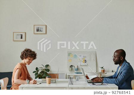 Businessman and businesswoman sitting at their workplaces opposite each other and working on computers at office Businessman and businesswoman sitting at their workplaces opposite each other and working on computers at office 92768541