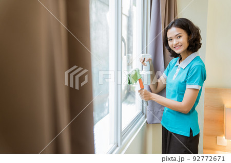 smiling female housekeeper in turquoise uniform wiping windows 92772671