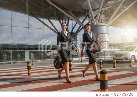 Happy stewardesses holding bags and crossing the road near terminal Happy stewardesses holding bags and crossing the road near terminal 92775720