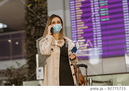 Female traveler in face mask carrying luggage bag at airport and talking phone before boarding 92775723