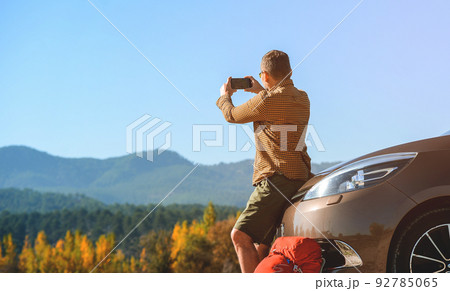 Man sits on car bumper using mobile phone to make picture of beautiful mountains view during summer trekking. Tourist with backpack stop to relax and enjoy nature Man sits on car bumper using mobile phone to make picture of beautiful mountains view during summer trekking. Tourist with backpack stop to relax and enjoy nature 92785065